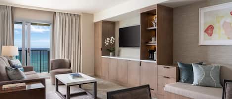 A modern hotel room with a balcony, a flat-screen TV, a sofa, a coffee table, and a seating area with cushions.