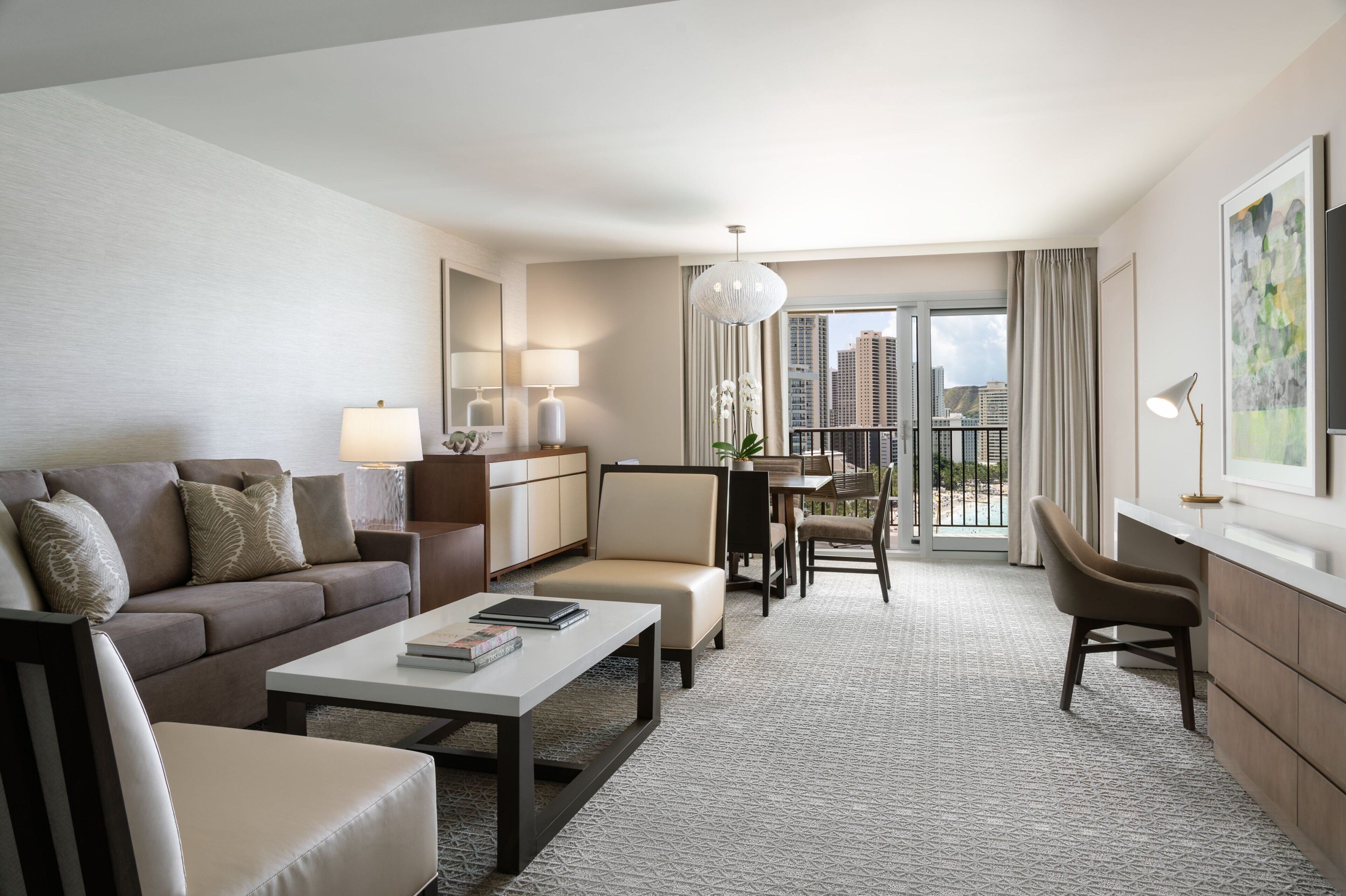 A modern hotel room with a sofa, a dining table, and chairs, a small desk, and a large window with a view of the city.