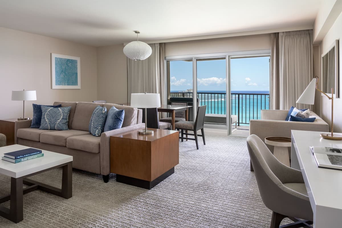 A modern hotel room with a sofa, a coffee table, a dining table, and a balcony with a view of the ocean.