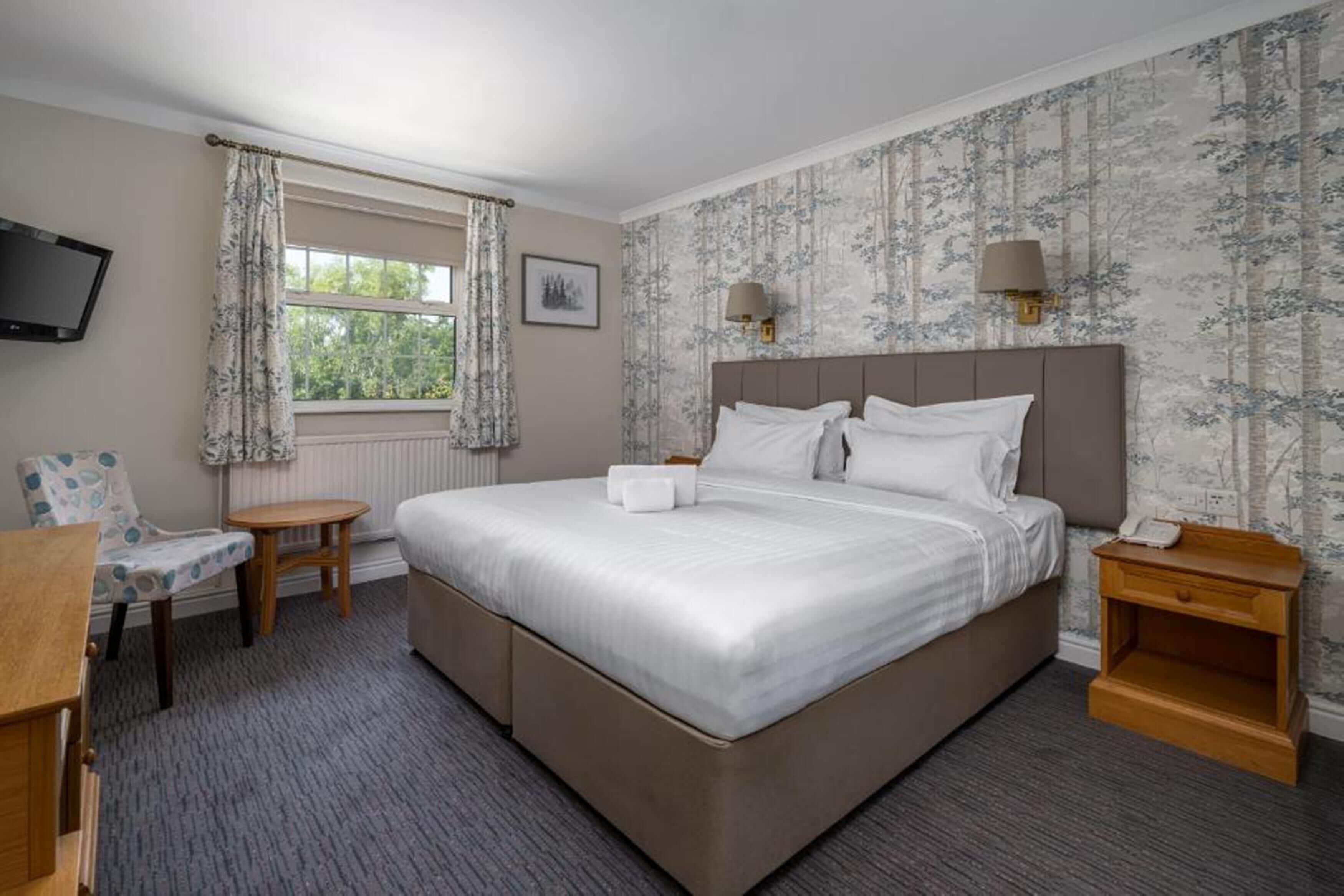 A bedroom with a large bed, a bedside table, a chair, a small table, a TV mounted on the wall, and a window with curtains.
