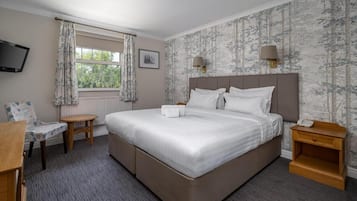 A bedroom with a large bed, a bedside table, a chair, a small table, a TV mounted on the wall, and a window with curtains.