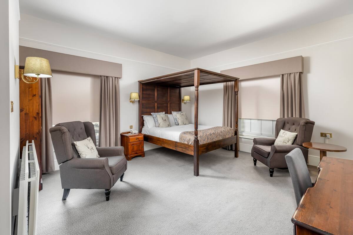 A four-poster bed with a wooden frame, two upholstered armchairs, a small wooden table, and a nightstand with a lamp.