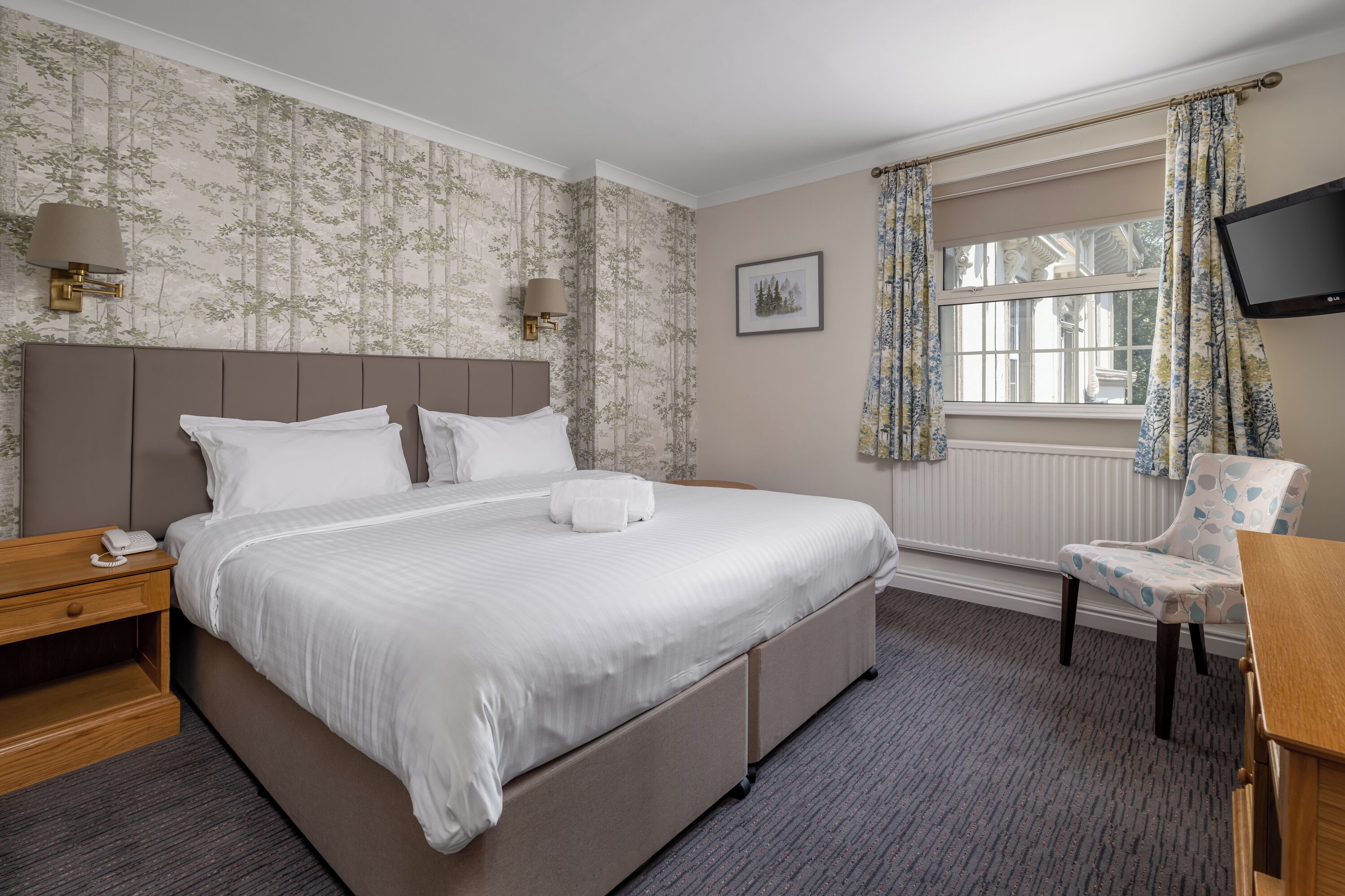Une chambre d’hôtel avec un grand lit, une table de chevet avec une lampe, une chaise, un téléviseur et une fenêtre avec des rideaux.