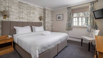 Une chambre d’hôtel avec un grand lit, une table de chevet avec une lampe, une chaise, un téléviseur et une fenêtre avec des rideaux.