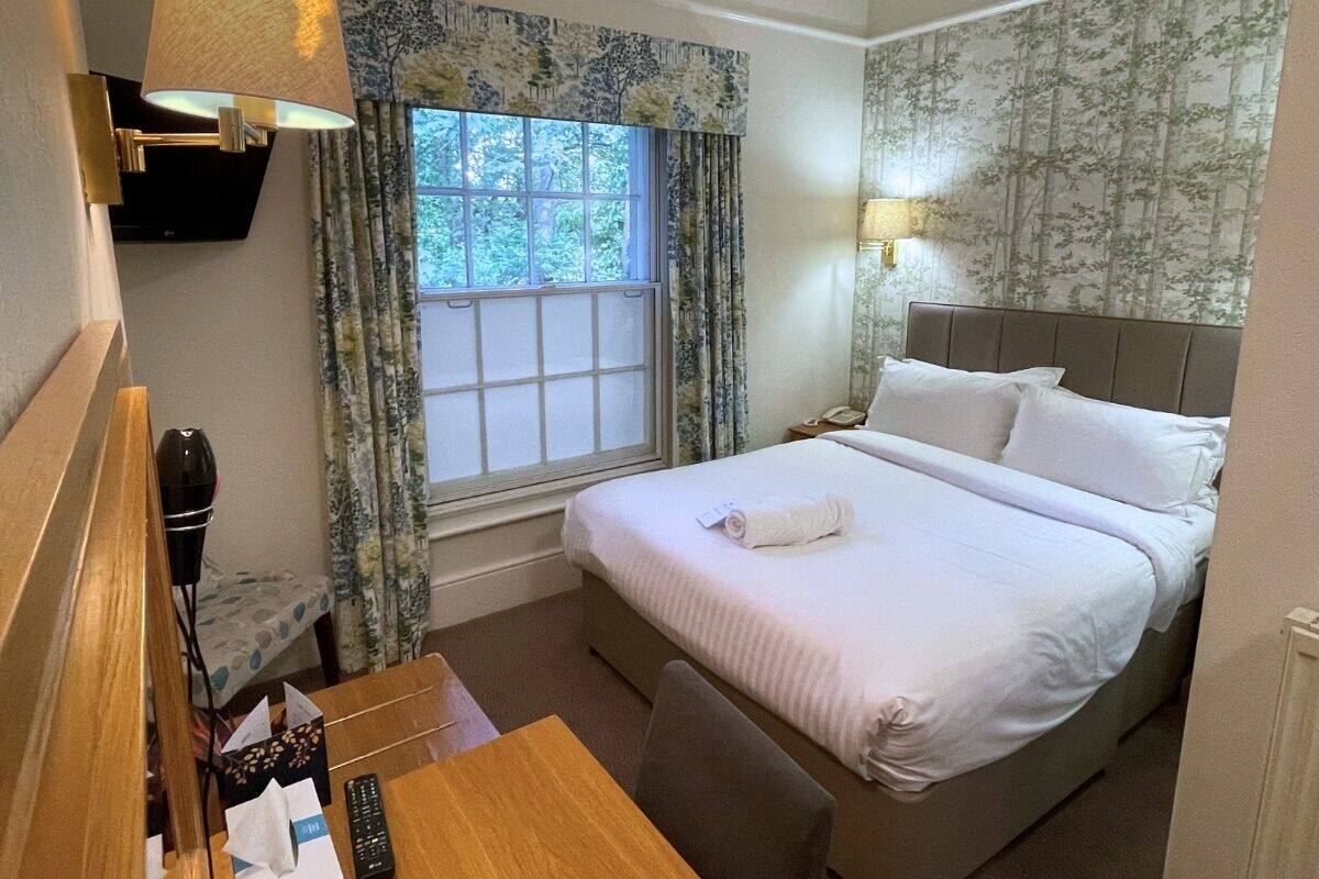 A hotel room with a bed, a desk, a chair, a window with curtains, and a wall-mounted lamp.