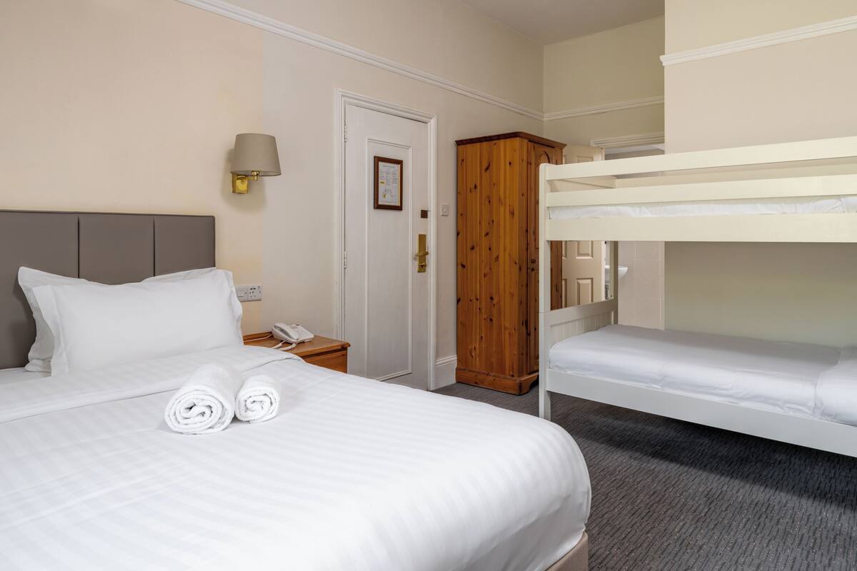 A hotel room with a single bed, a bunk bed, a wooden wardrobe, a bedside table with a lamp, and a door with a notice.