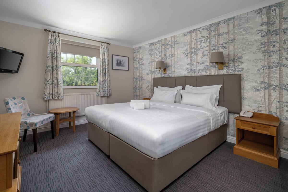 A hotel room with a large bed, a small table, a chair, a bedside table with a lamp, and a TV mounted on the wall.