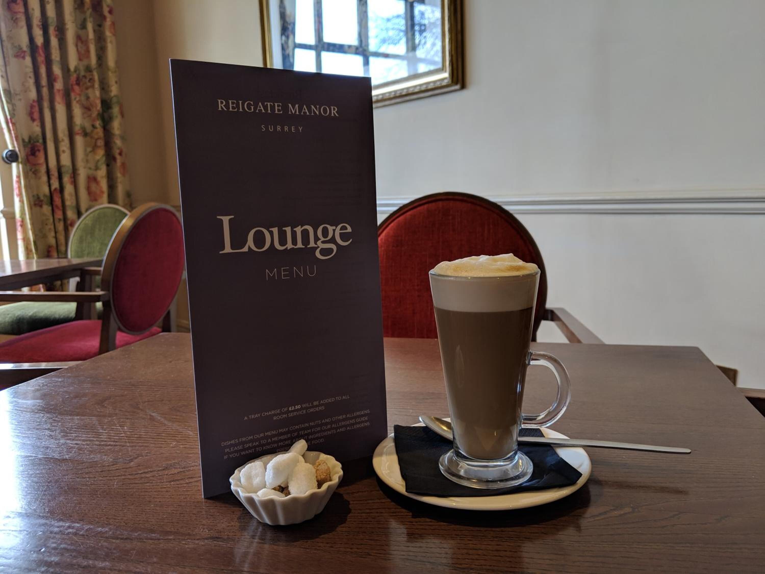 Quadro de menu intitulado 'Reigate Manor Lounge Menu', com aviso de taxa de bandeja, um copo alto de café com espuma e uma pequena tigela de cubos de açúcar sobre uma mesa de madeira.