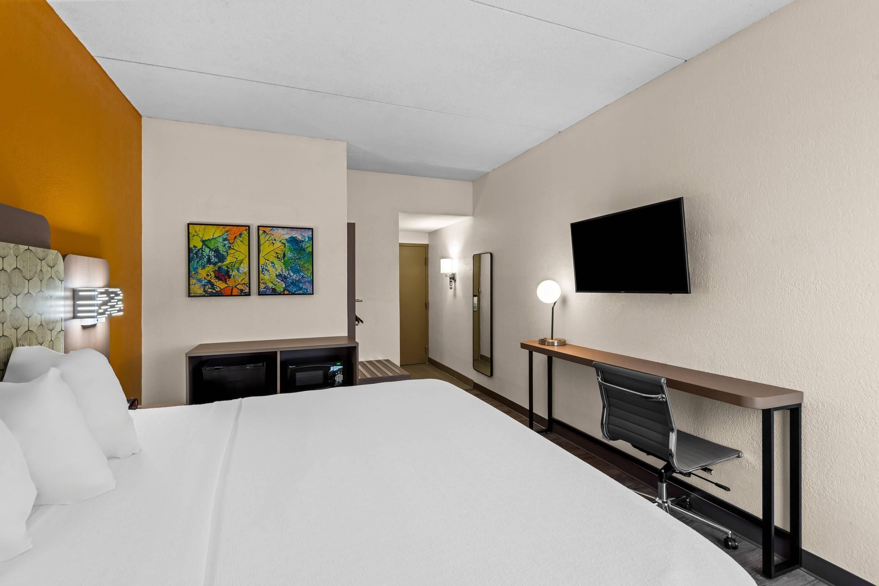 A hotel room with a bed, desk, chair, TV, artwork, and a lamp.