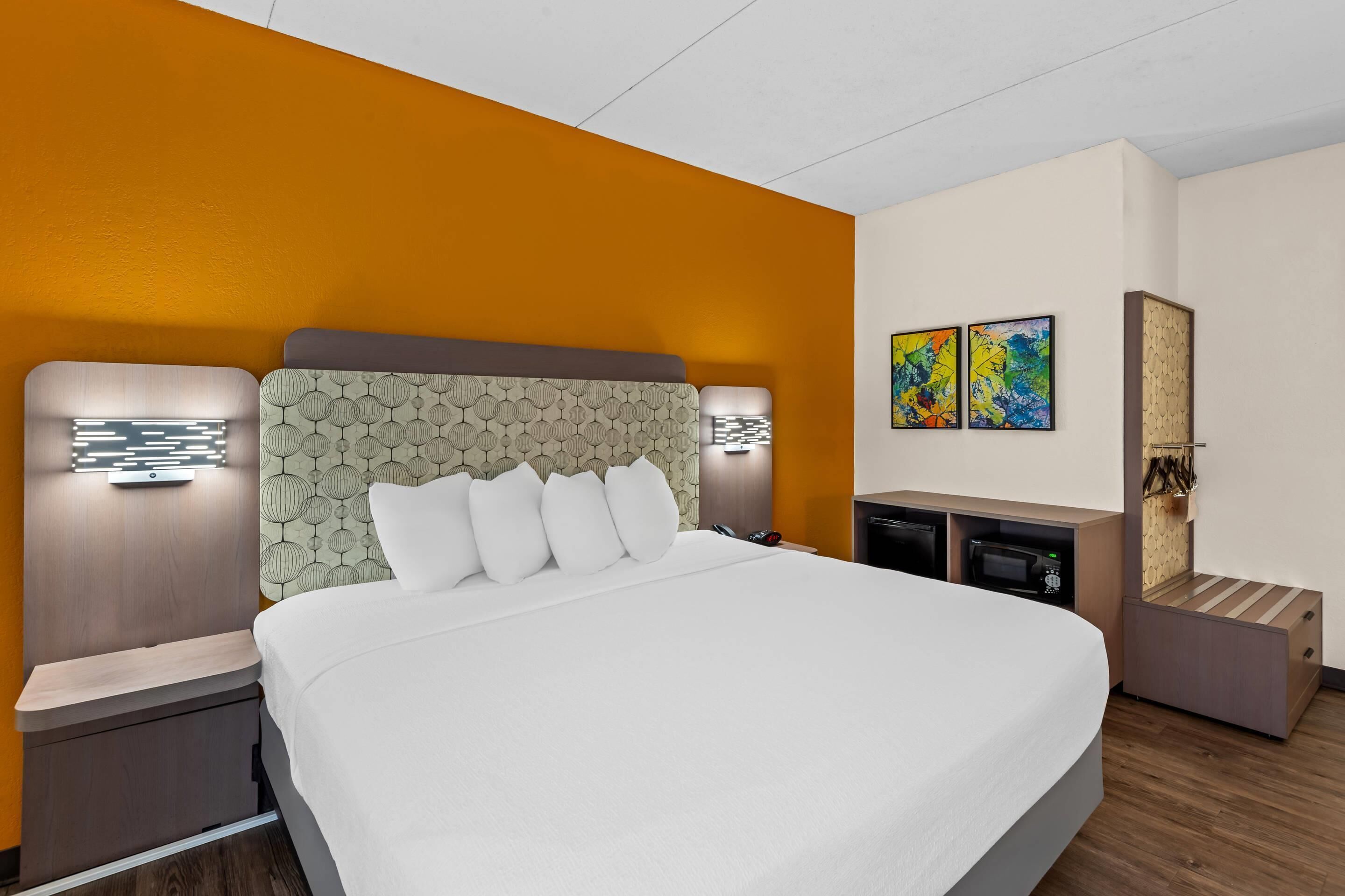 A modern hotel room with a large bed, a wooden nightstand, a desk with a chair, and a small seating area.