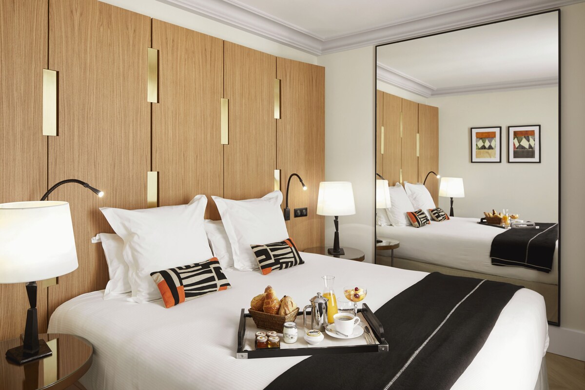 A hotel room with a large bed, bedside lamps, a tray with breakfast items, and a mirror reflecting the room.