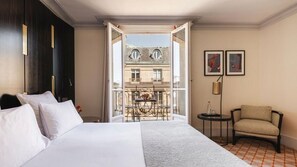 A hotel room with a bed, a chair, a small table, and a balcony with a view of buildings. - Hotel Montalembert (Paris)