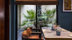 A dining area with a table set for two, a window with a view of a garden, and a large plant in the background. - Hotel Montalembert (Paris)