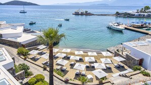 Un hotel frente al mar con zona de playa, tumbonas y sombrillas, un puerto deportivo con barcos y montañas al fondo.