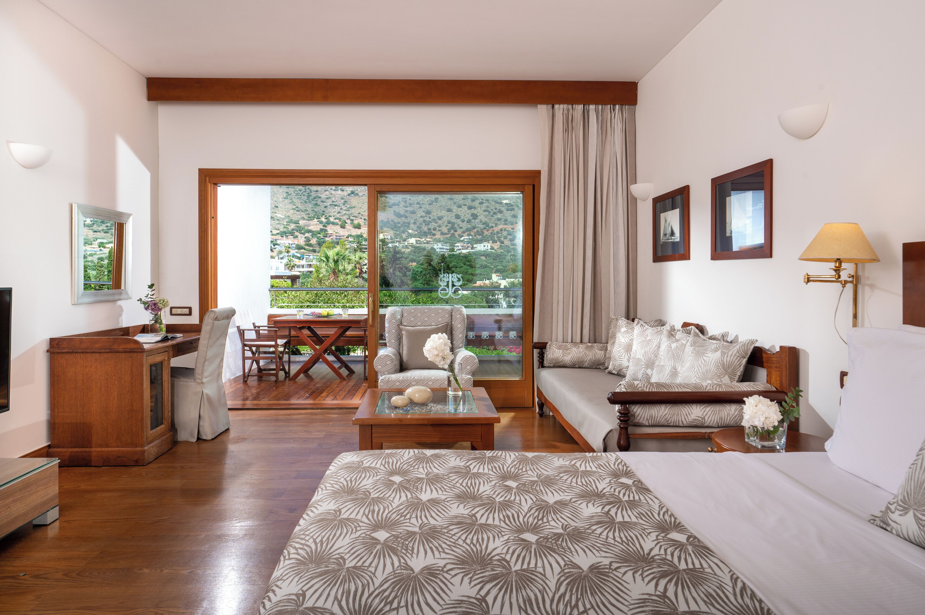 A hotel room with a large bed, a sofa, a dining area, and a balcony with a view of the hills.