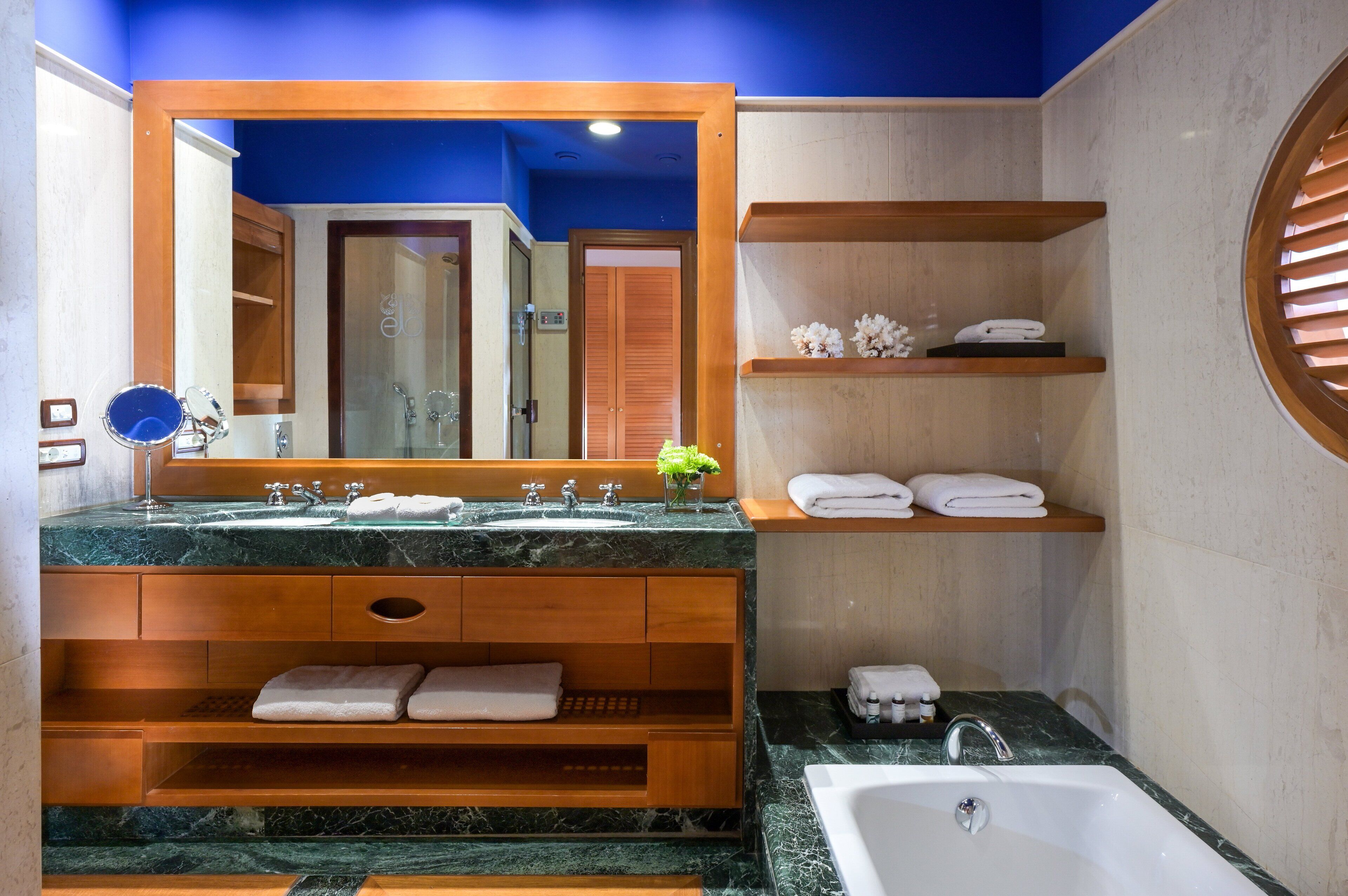 Un baño moderno con un espejo grande, encimeras de mármol, tocador de madera, repisas con toallas y una bañera.