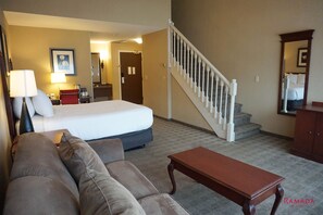 A hotel room with a bed, a sofa, a wooden table, a staircase, and a bathroom visible in the background. - Ramada Hotel & Conference Center by Wyndham Kelowna (Kelowna)