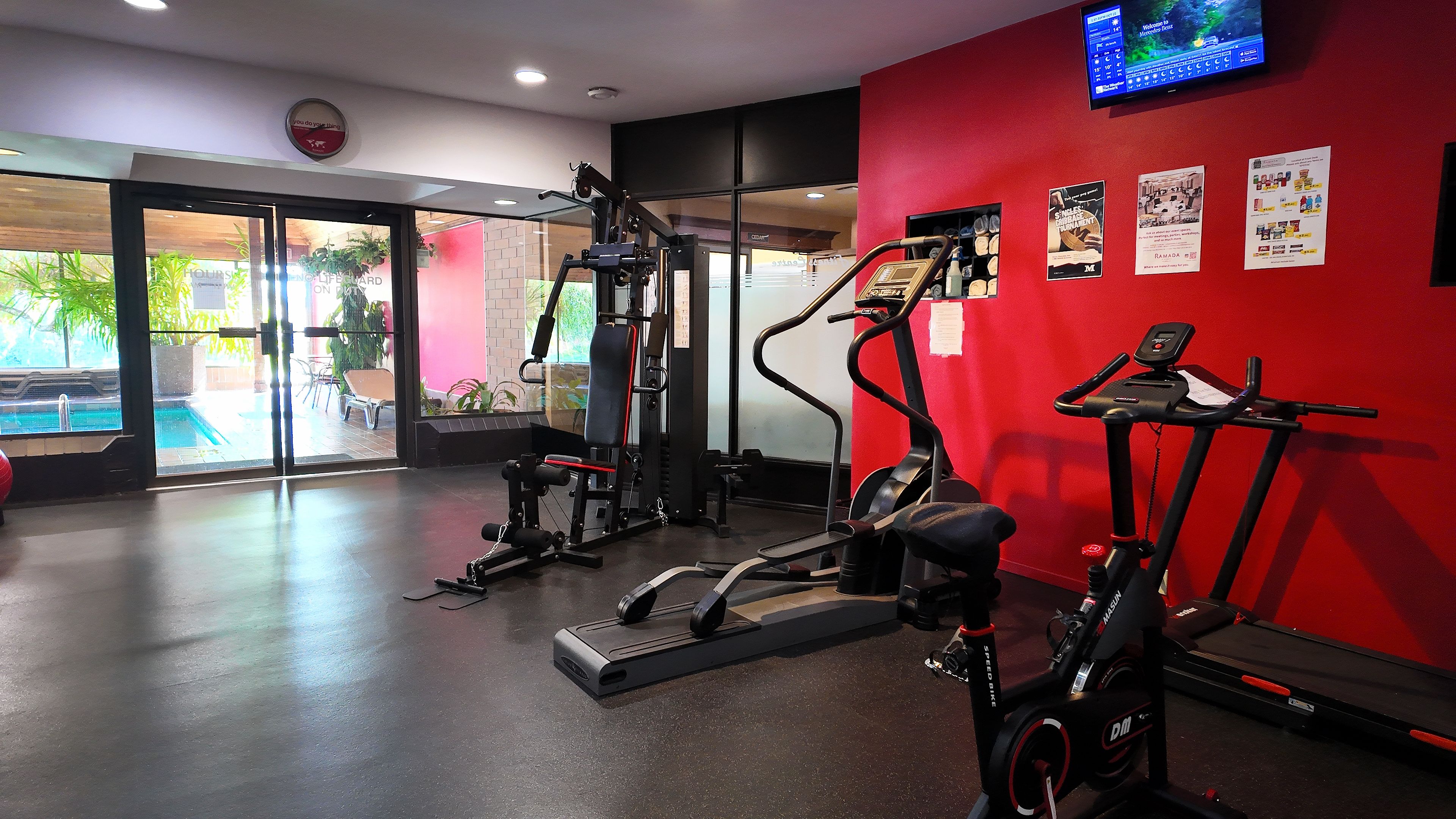 Un gimnasio con equipo de ejercicio, un televisor y una piscina visibles a través de puertas de vidrio.