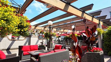 Un área de estar exterior con mobiliario de mimbre, cojines rojos y plantas en macetas.