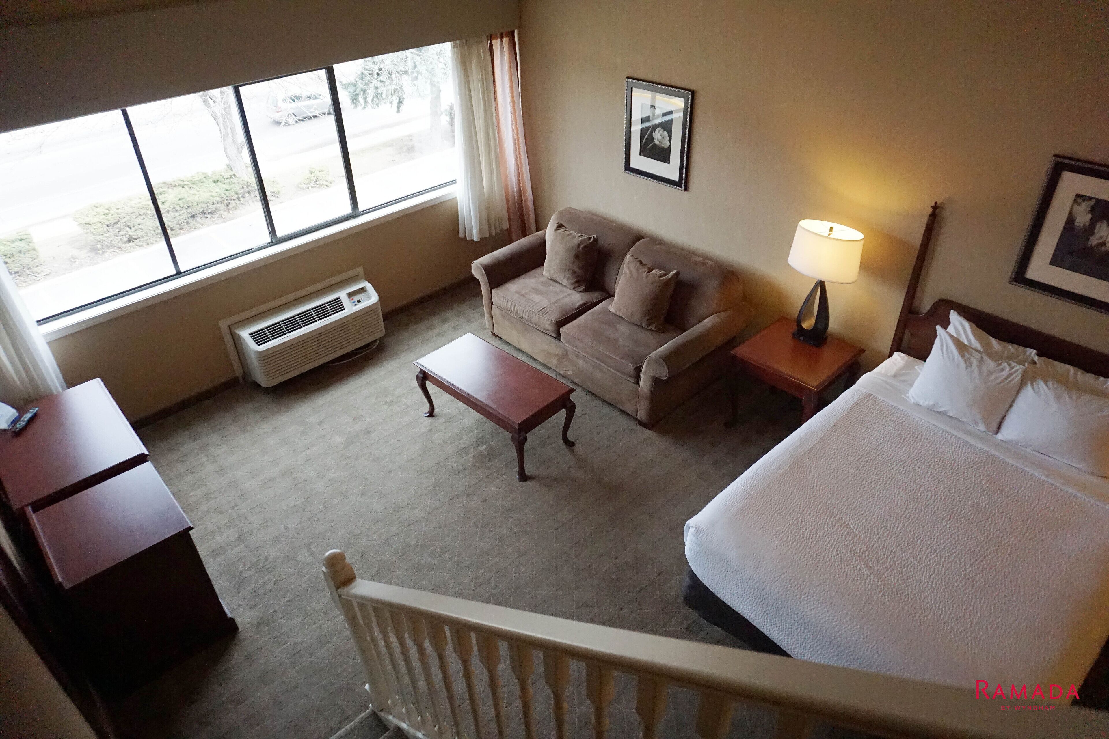 Une chambre d’hôtel comprenant un lit, un canapé, une petite table, une table de chevet avec une lampe, une fenêtre avec des rideaux et un tableau encadré au mur.
