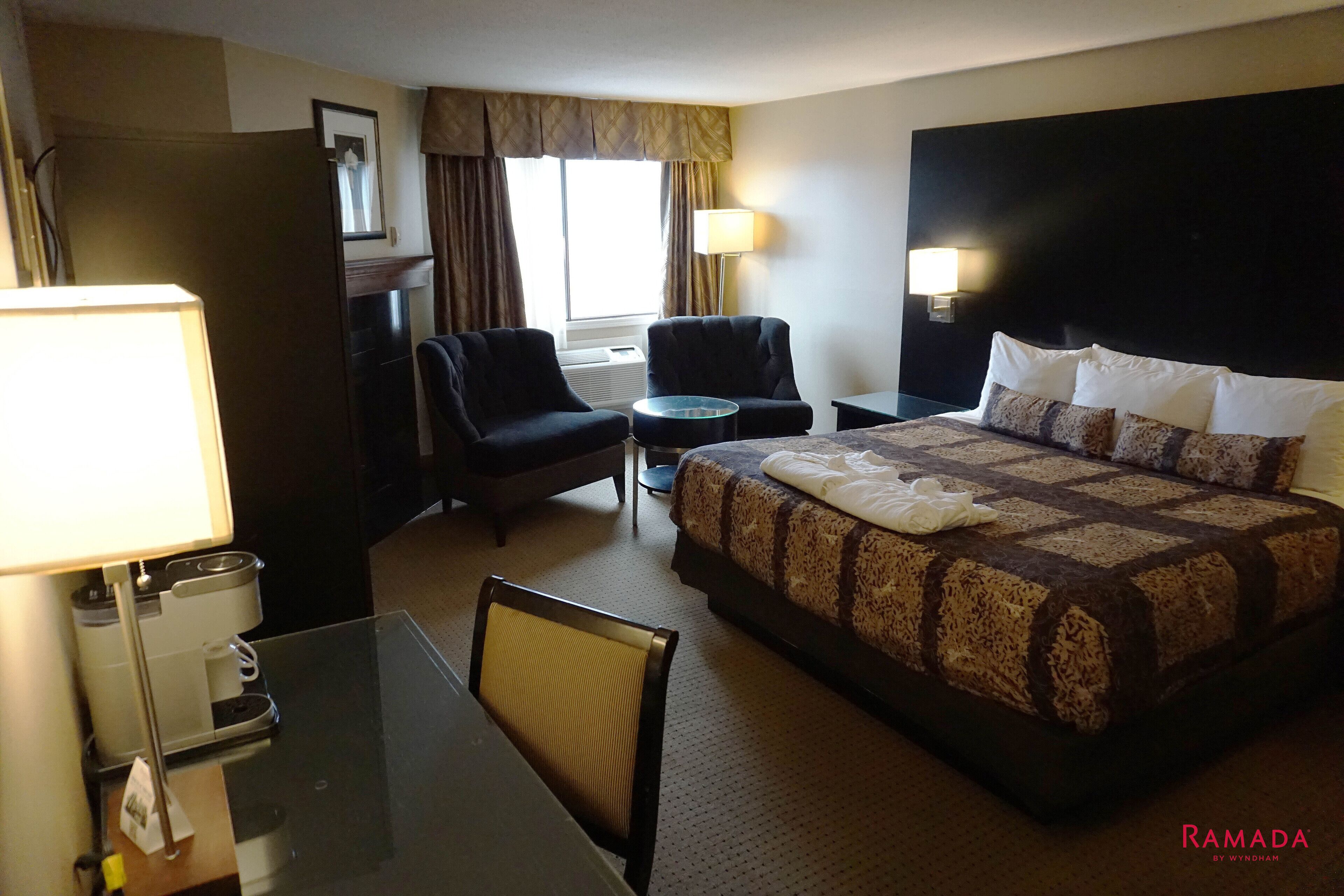 A hotel room with a large bed, two armchairs, a small table, a desk with a lamp, and a coffee machine.