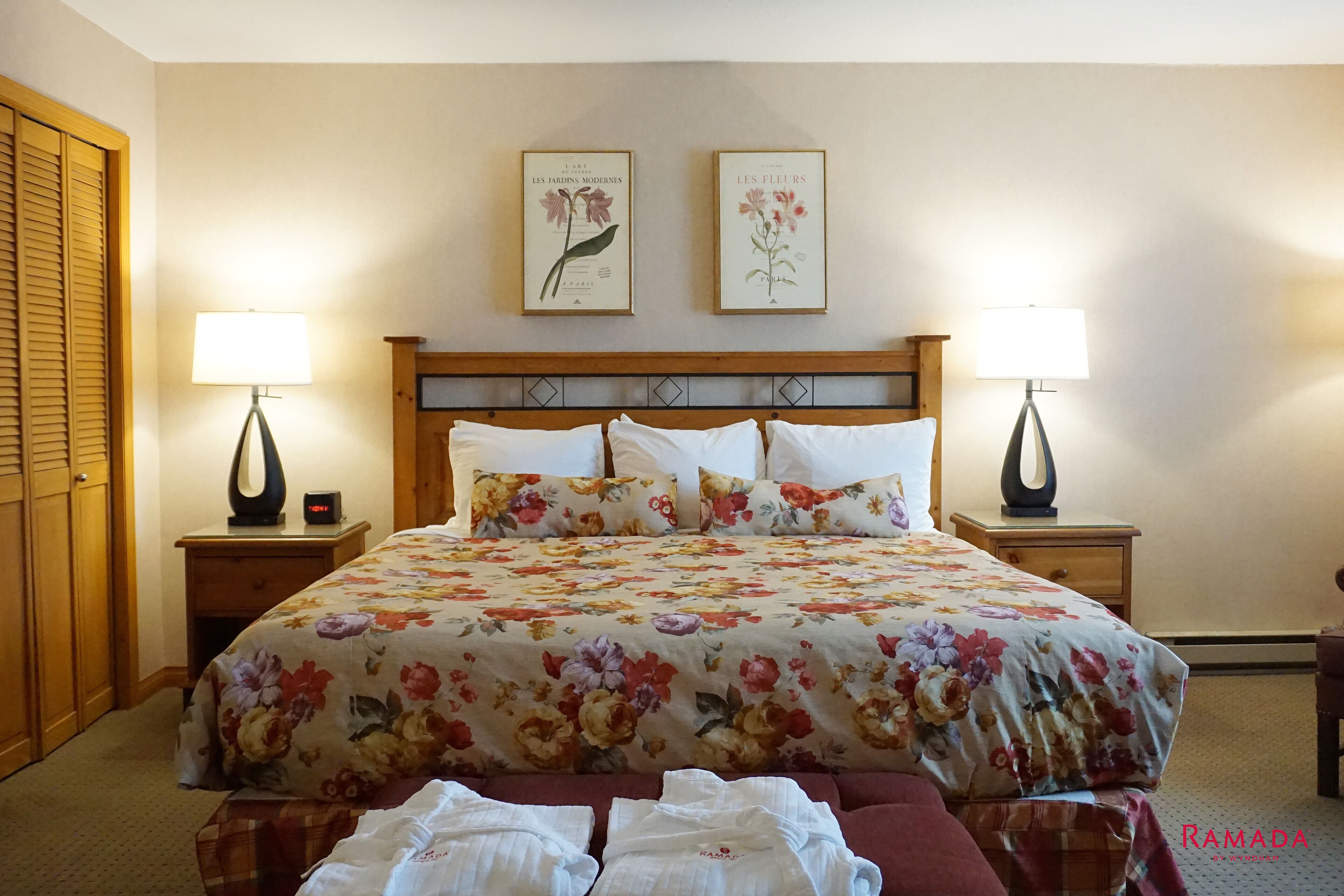 A hotel room with a floral bedspread, two wall-mounted lamps, and two framed botanical prints above the bed.