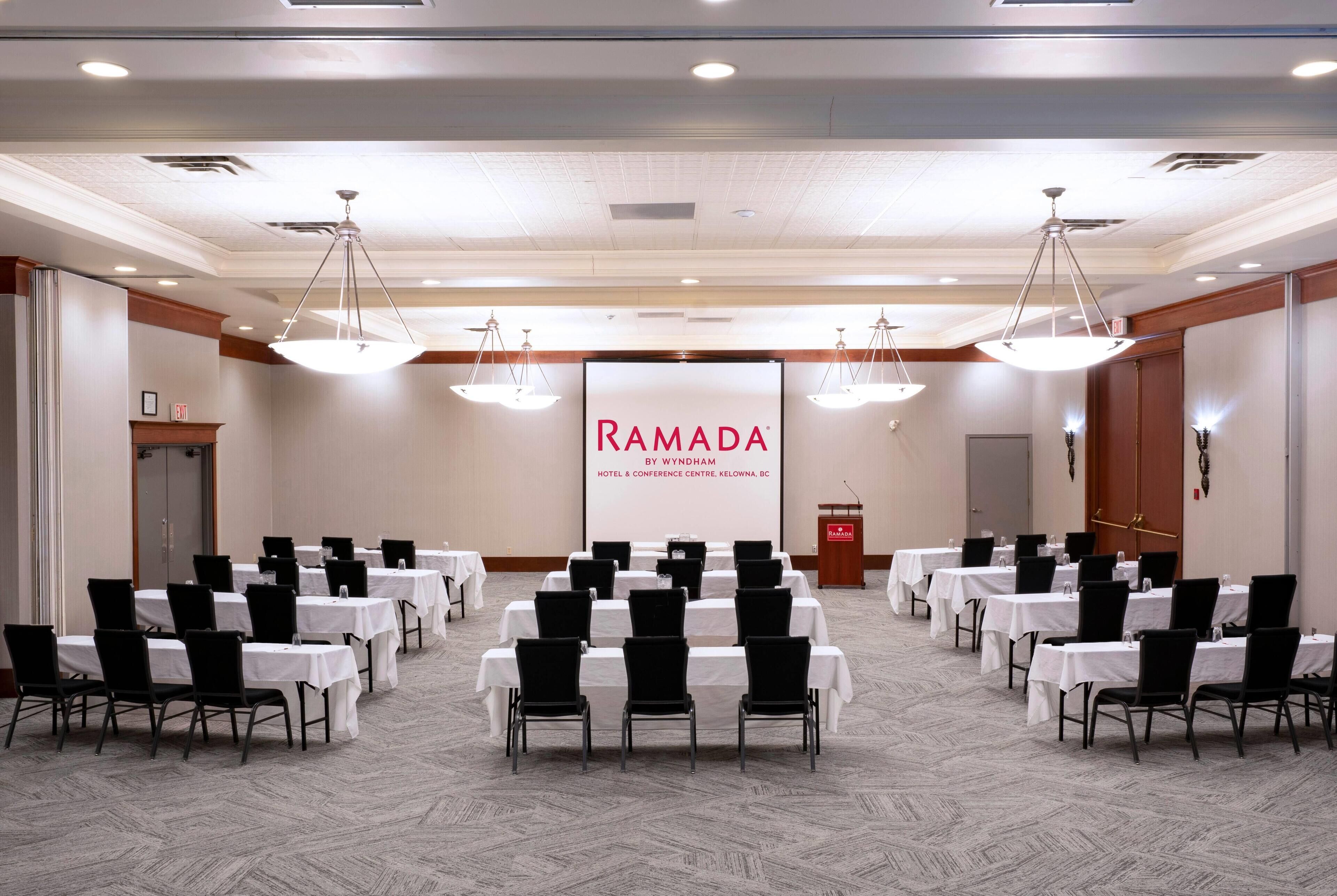 Un salón de conferencias con filas de mesas y sillas, una pantalla grande mostrando el nombre del hotel y un atril.