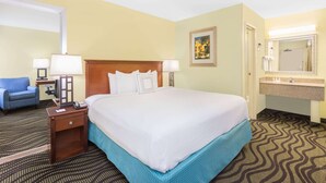 Habitación de hotel con una cama grande, un escritorio con lámpara, una silla, un baño con lavamanos y espejo, y un cuadro en la pared.