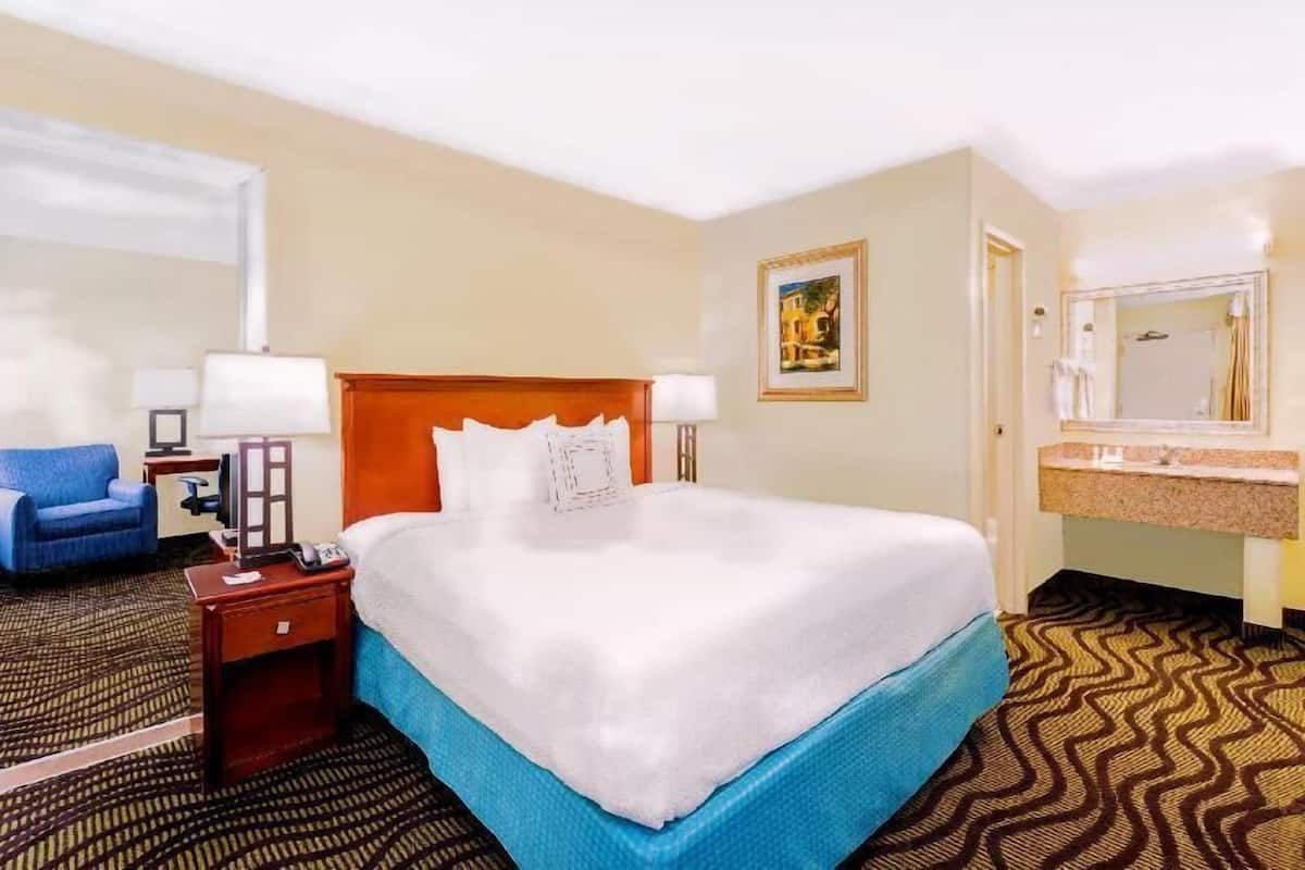 A hotel room with a bed, a desk with a lamp, a chair, a bathroom with a sink and mirror, and a patterned carpet.