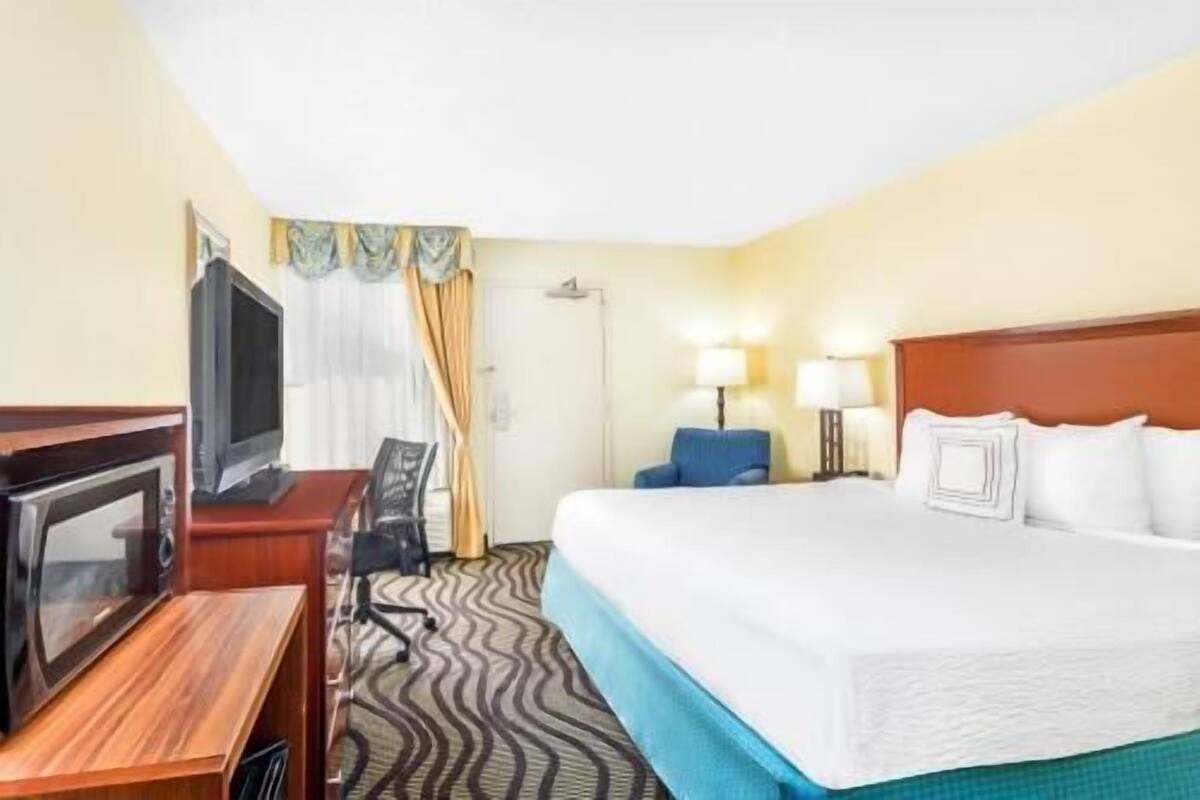A hotel room with a bed, a desk with a television, a chair, a lamp, and a patterned carpet.