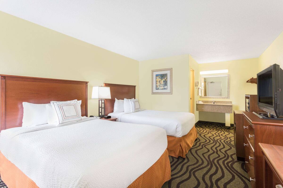 A hotel room with two beds, a television, a desk with a lamp, a mirror, and a bathroom with a sink and mirror.