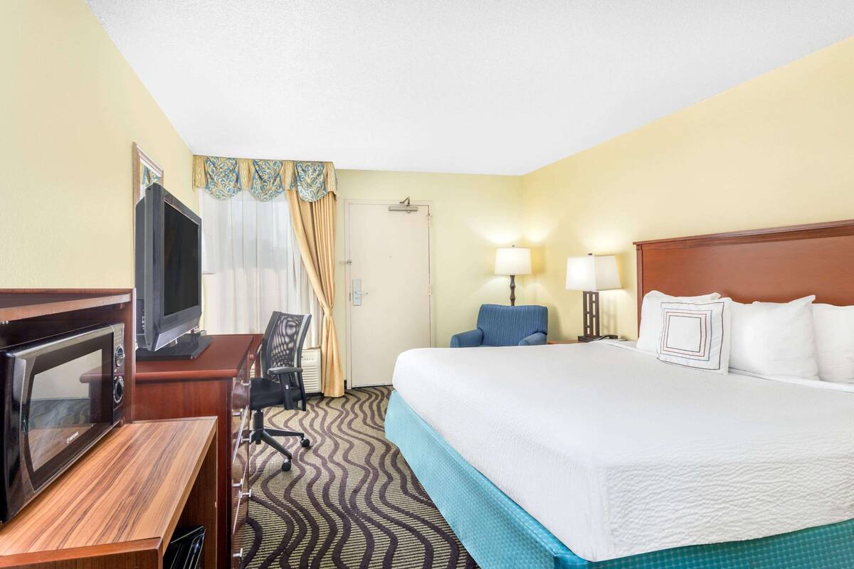 A hotel room with a bed, a desk with a microwave, a television, a chair, and a lamp.