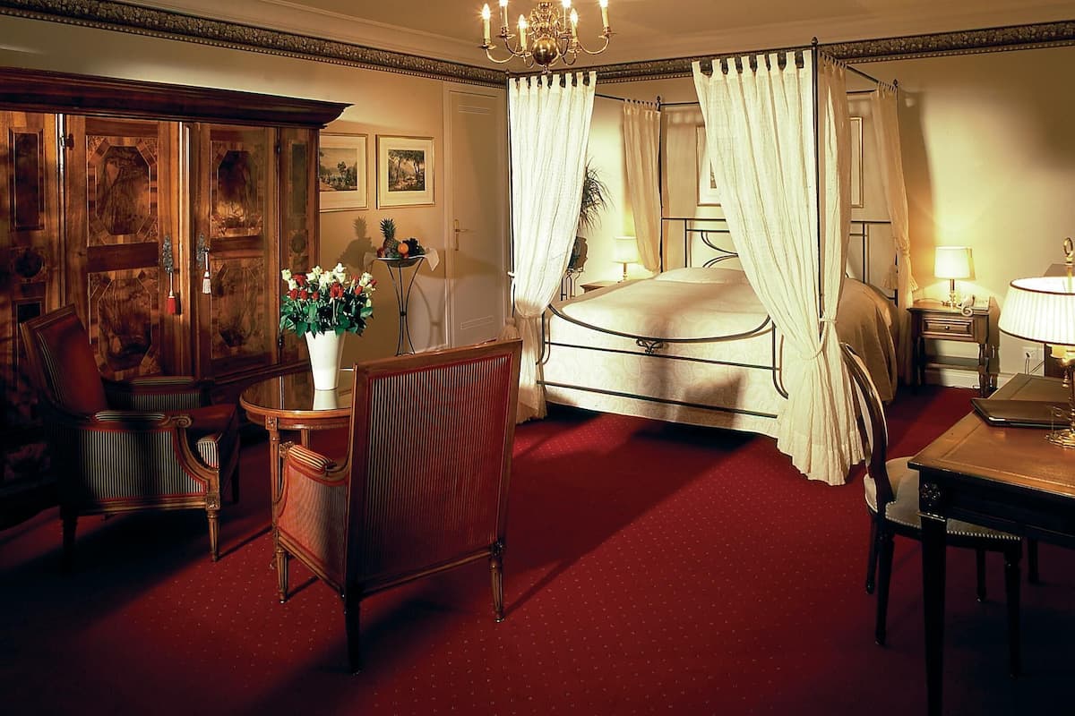 A spacious bedroom with a large bed, a sitting area with a chair and table, and a chandelier.