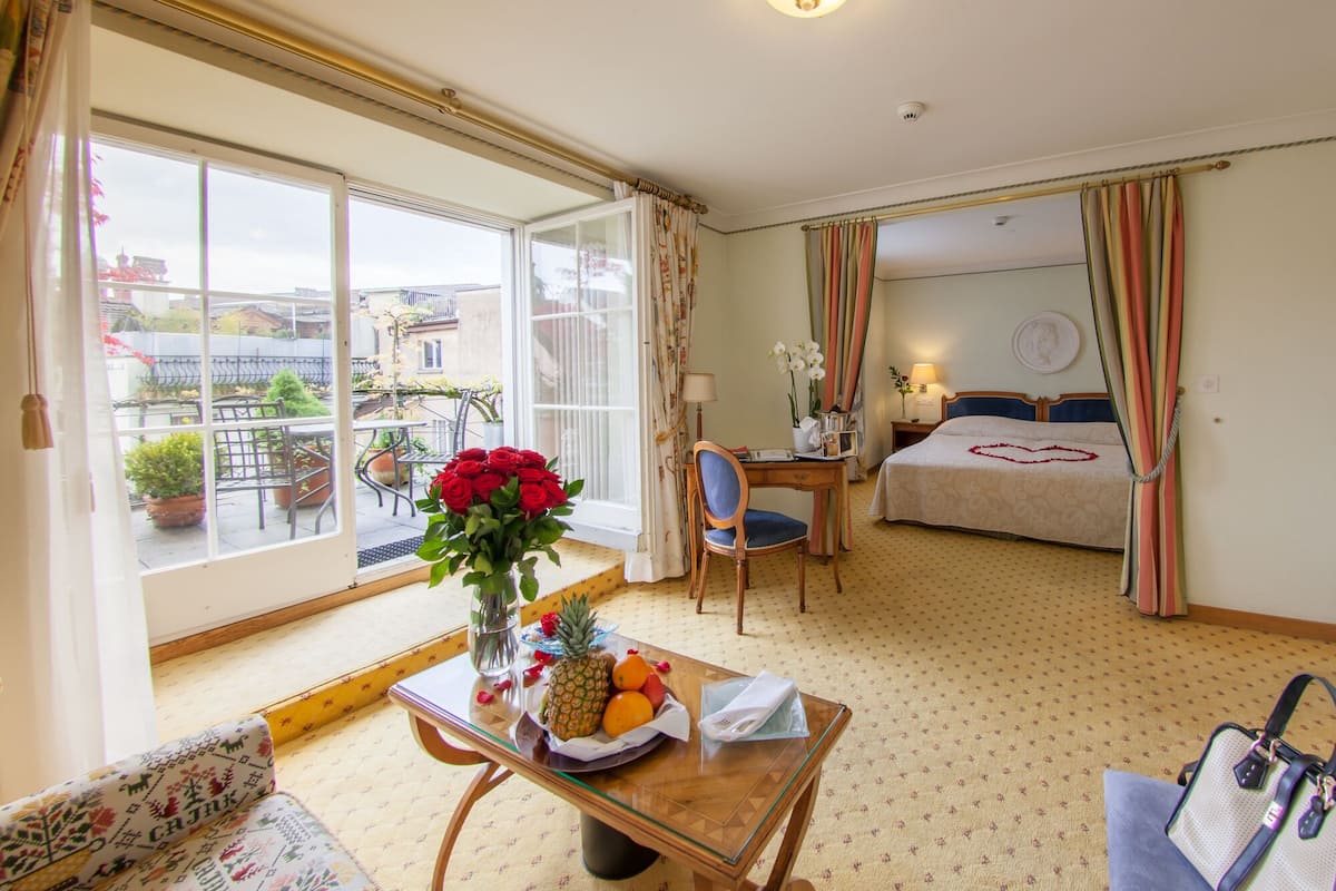 A hotel room with a large window, a bed with a heart-shaped decoration, a desk with a lamp, and a table with fruits and flowers.