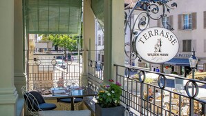 Une terrasse avec des places assises extérieures, une enseigne indiquant « Terrasse Wilden Mann », et une vue sur la rue et les bâtiments.