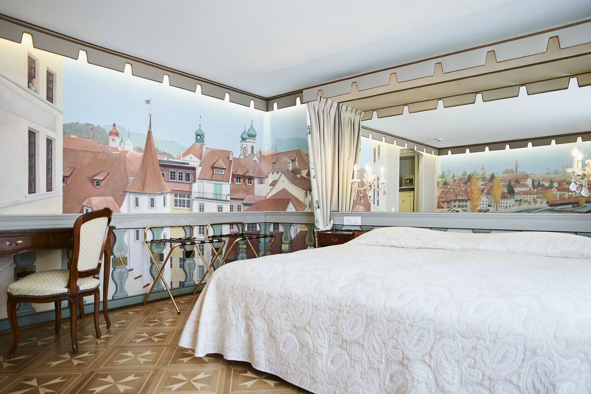 A hotel room with a bed, a desk, a chair, and a view of a cityscape through a large window.