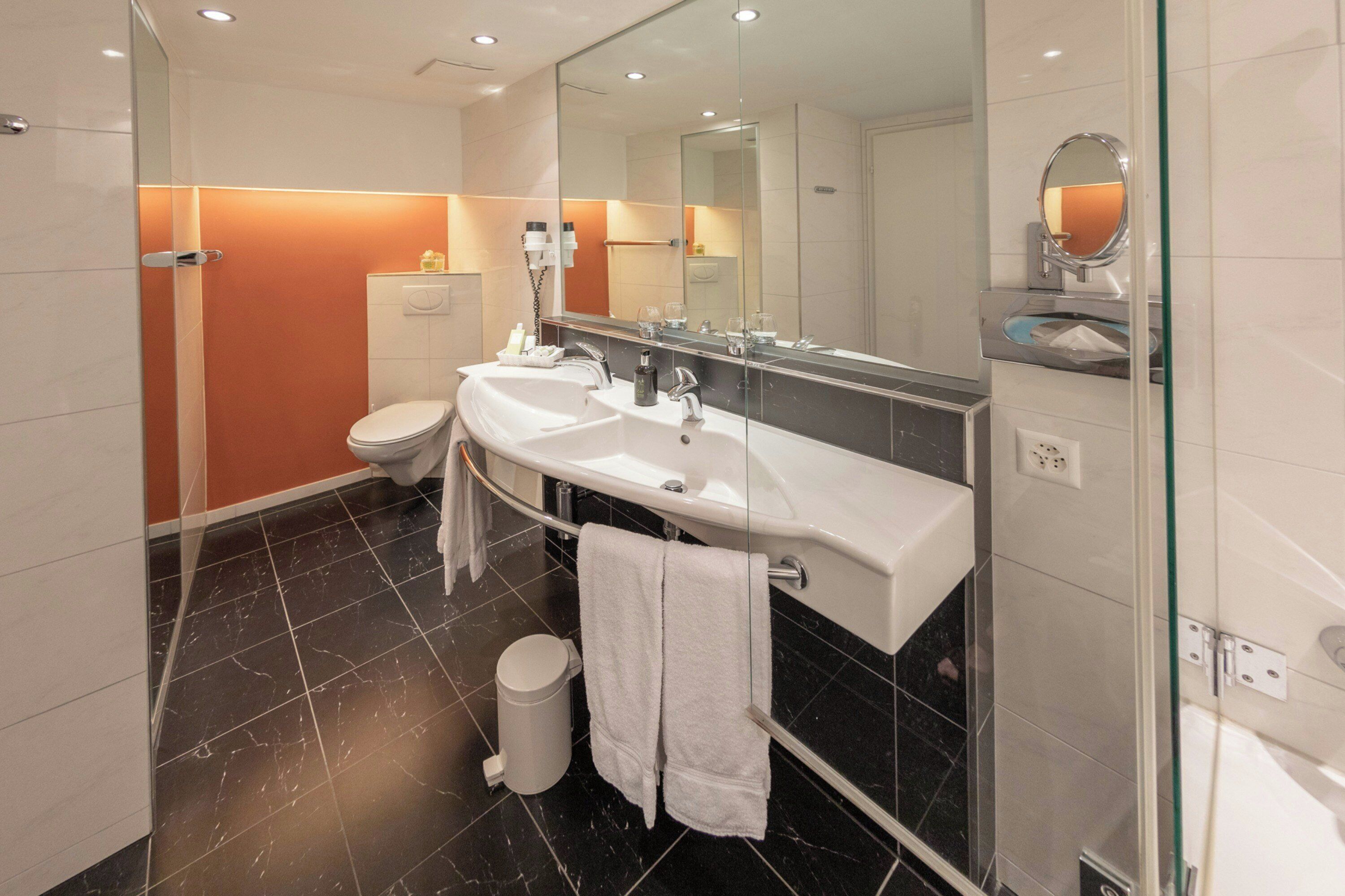 A modern bathroom with a glass shower enclosure, a white sink with a large mirror, and a black-tiled floor.