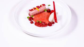 Ein weißer Teller mit einem Dessert bestehend aus einem zylindrischen, rosa farbenen Element, roten Beeren und einer weißen Kugel, begleitet von einem roten Besteckstück.