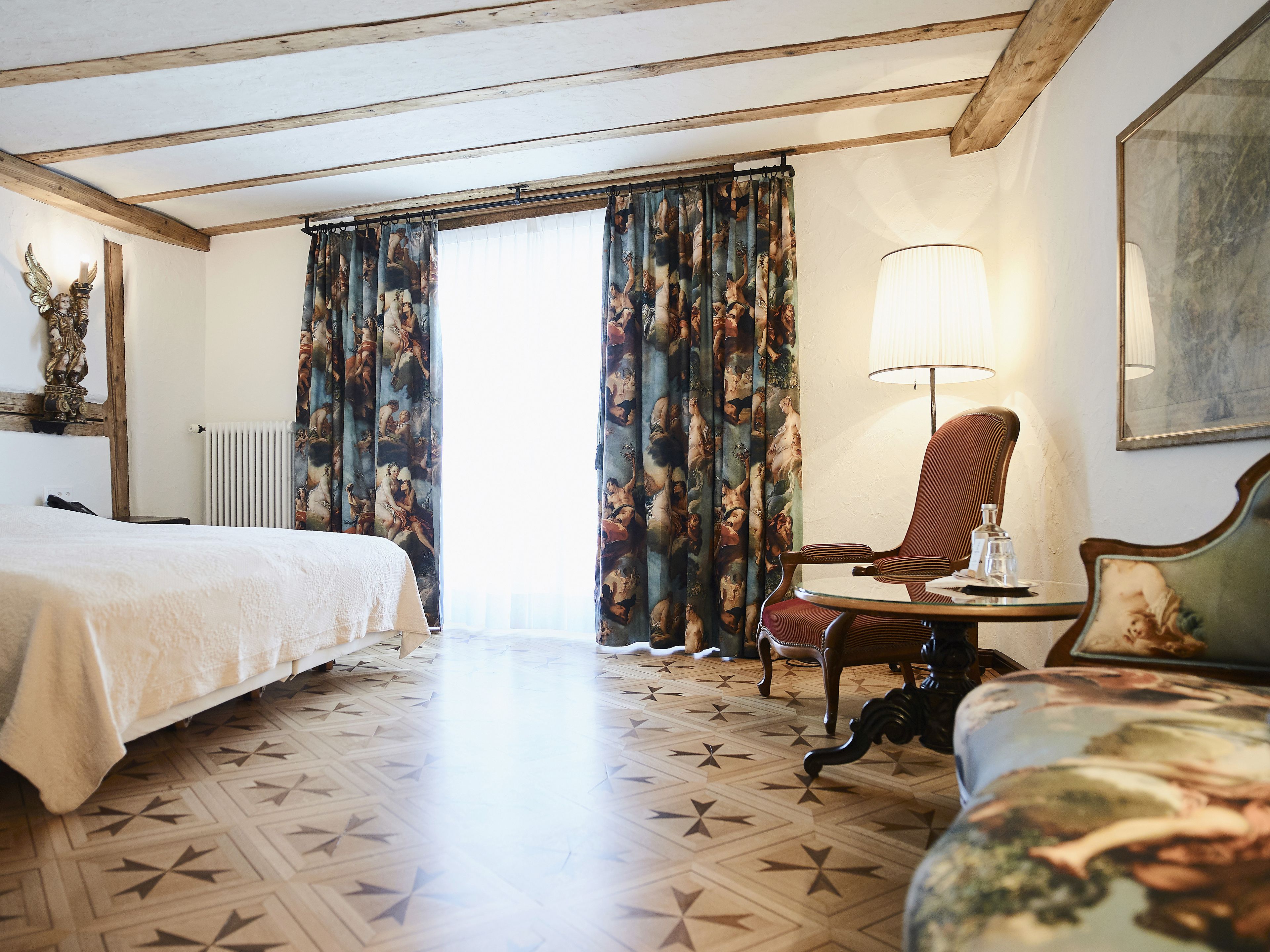 Una camera d'albergo con un letto, una sedia, un tavolo con una lampada e un'ampia finestra con tende.