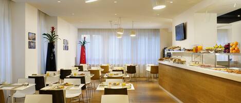 A hotel dining area with tables set for breakfast, a buffet with various food items, and a large window with curtains.