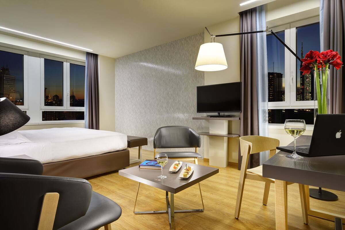 A hotel room with a bed, a desk with a laptop, a chair, a dining table with food and a glass of wine, and a TV.