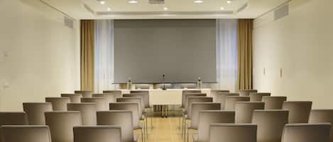 Un salón de conferencias con filas de sillas, una pantalla de proyector y una mesa con micrófonos.