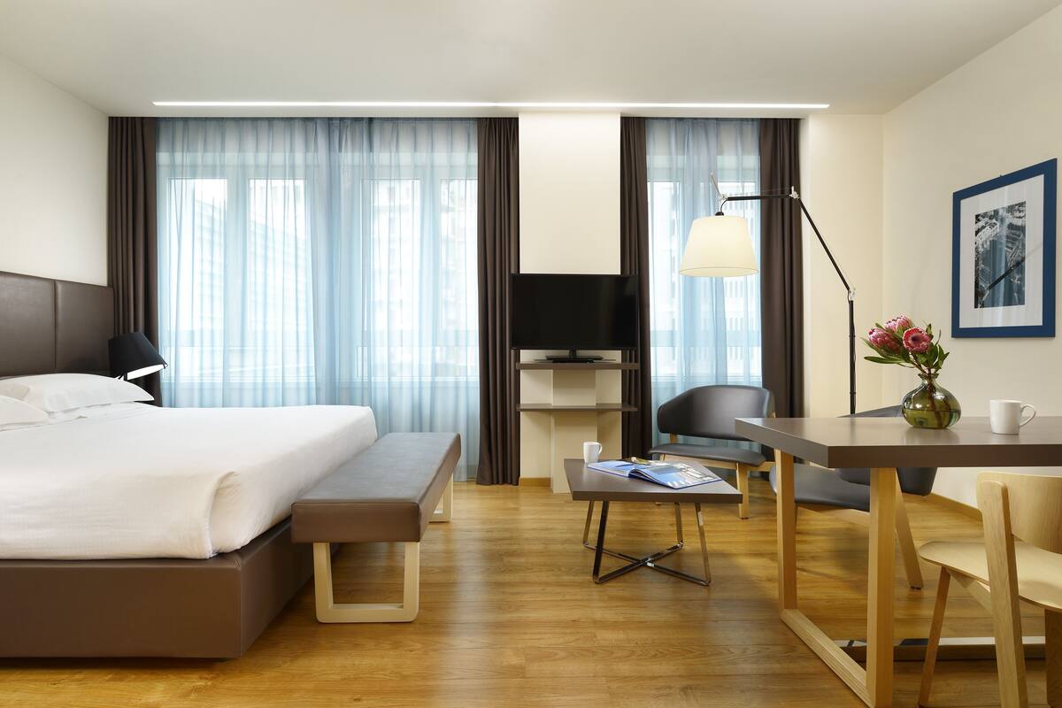 A modern hotel room with a bed, desk, chair, and a small table with a vase and a cup.
