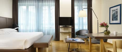 A modern hotel room with a bed, desk, chair, and a small table with a vase and a cup.