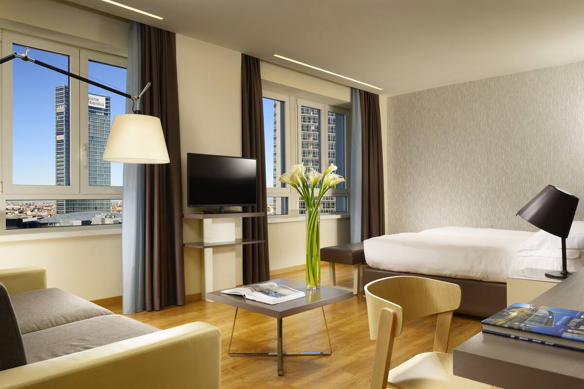 A modern hotel room with a bed, a TV, a desk with a lamp, a chair, and a view of the cityscape.