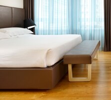 A modern hotel room with a large bed, a bedside table, a lamp, and a view of the outdoors through sheer curtains.