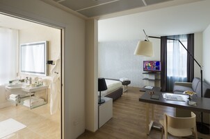 A modern hotel room with a bathroom, a bed, a desk, and a television.