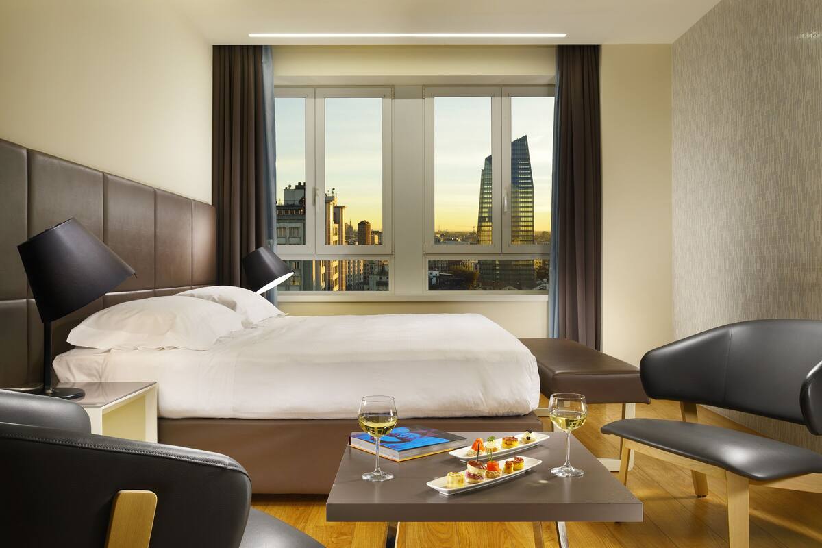 A hotel room with a bed, a bedside table with a lamp, a coffee table with a wine glass and a plate of food, and a view of the city skyline through large windows.