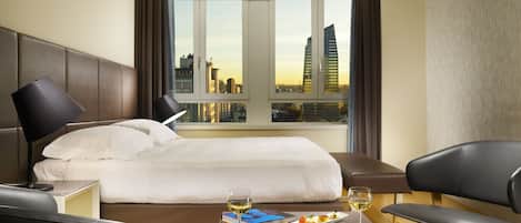 A hotel room with a bed, a bedside table with a lamp, a coffee table with a wine glass and a plate of food, and a view of the city skyline through large windows.