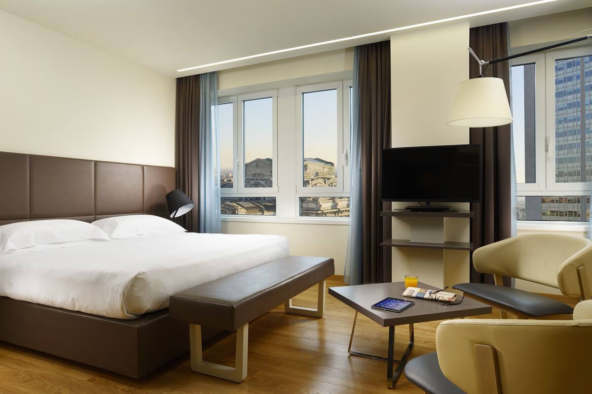 A hotel room with a bed, a bench, a table with a book and a glass of orange juice, a TV, and a chair.